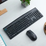 Black keyboard with Thai-English characters and a black mouse on a light gray desk. A small plant in a pot is nearby, enhancing the office setup.