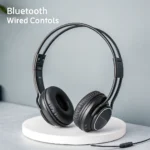 Black wireless headphones with Bluetooth and wired controls displayed on a white circular platform against a gray background.