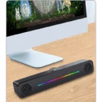 Black desktop soundbar with a rainbow light on wooden desk in front of a computer monitor.