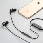 Black wired earbuds with control module next to a gold-colored smartphone on a light surface.