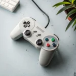A gray, retro-style wired game controller with colorful buttons lies on a light blue desk next to a keyboard and a potted plant.