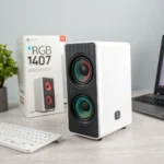 White RGB 1407 speaker with colorful dual drivers on a desk with a keyboard, laptop, and plant, product box in background.