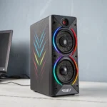 Black gaming speaker with dual woofers and RGB lighting, placed on a light-colored desk next to a computer monitor.