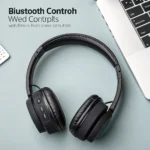 Black wireless headphones with control buttons placed beside a laptop with visible keyboard, on a light-colored desk.